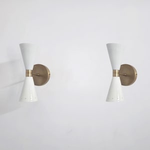 Peut inclure: Deux appliques murales blanches avec un centre et une plaque arrière de couleur laiton. Chaque applique a une forme conique, avec un sommet plus large et un bas plus étroit. Les appliques sont montées sur un mur blanc.