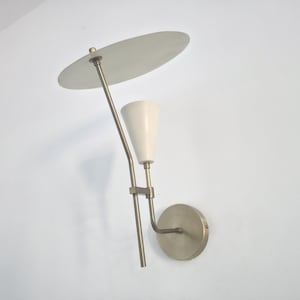 Op de afbeelding: Een wandlamp in mid-century modern stijl met een witte conische kap en een cirkelvormige, gebroken witte schijfkap. De armatuur heeft een messing afwerking en is gemonteerd op een cirkelvormige basis.