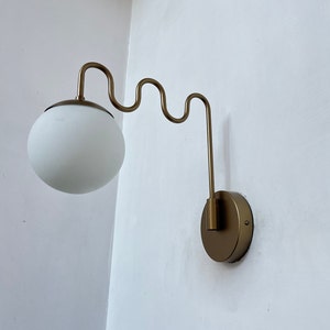 Pode incluir: Uma arandela de parede dourada com um abajur de vidro branco. A arandela tem um braço curvo que se estende da parede.