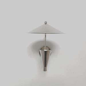 May include: A modern, silver-toned wall sconce with a conical shade and a small, round finial. The lamp has a minimalist design, with a sleek, metallic finish. The sconce is mounted on a white wall, creating a clean, contemporary aesthetic.
