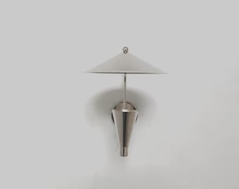 May include: A modern, silver-toned wall sconce with a conical shade and a small, round finial. The lamp has a minimalist design, with a sleek, metallic finish. The sconce is mounted on a white wall, creating a clean, contemporary aesthetic.