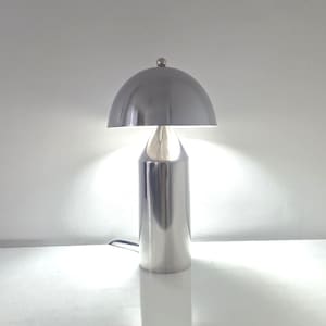 Puede incluir: Una lámpara de mesa con acabado en cromo pulido que presenta una base cilíndrica y una pantalla en forma de cúpula. La lámpara emite una luz blanca brillante desde debajo de la pantalla, creando un resplandor contra la pared. Un remate esférico corona la cúpula.