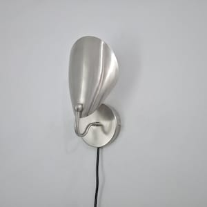 Pode incluir: Arandela de parede em níquel escovado com uma sombra curva em forma de pétala. A luminária tem uma base circular e um braço curvo. Um cabo preto se estende da parte inferior. Montada em uma parede branca.