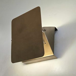 Pode incluir: Um candeeiro de parede com uma placa metálica retangular castanha mate posicionada à frente de uma base de metal dourado. O design inclui um mecanismo de dobradiça visível na lateral, permitindo ajustar a placa frontal para a luz.