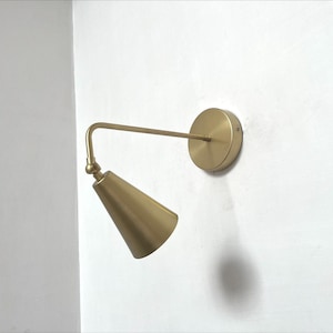 May include: A brass-colored wall-mounted sconce with a conical shade and a circular base. The lamp extends from the wall with a straight arm and a curved joint, creating a modern, minimalist design.