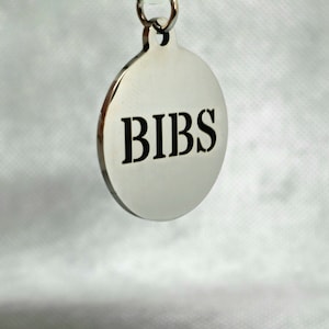 May include: A silver round tag with the word "BIBS" engraved in black lettering.