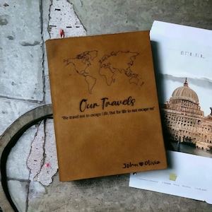 Puede incluir: Un diario de cuero marrón con un mapa del mundo grabado y el texto "Our Travels" y "We travel not to escape life, but for life to not escape us". El diario también tiene los nombres "John" y "Olivia" grabados en la parte inferior.