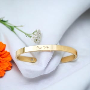 Könnte beinhalten: Ein goldfarbenes Armkettchen mit der Gravur "Para Vida" auf der Vorderseite.