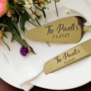 Può includere: Set di coltello e paletta per torta color oro con l'iscrizione "The Powell's 7.6.2025". Il set è posto su un piatto bianco con un tovagliolo bianco e una composizione floreale di rose rosa e verde.