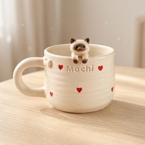 Puede incluir: Una taza de cerámica de color crema con una pequeña figura esculpida de un gato siamés posada en el borde. El exterior está decorado con pequeños corazones rojos en relieve y la palabra Mochi grabada en el centro. Tiene un asa redondeada.