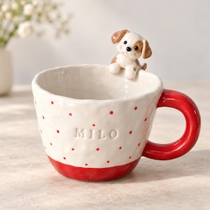 Könnte beinhalten: Ein weißer Keramikbecher mit strukturierter Oberfläche, roten Punkten und einem einfarbig roten Henkel. Der Name MILO ist auf der Vorderseite eingeprägt. Eine kleine, dreidimensionale Keramik-Welpenfigur ist am inneren Rand befestigt.