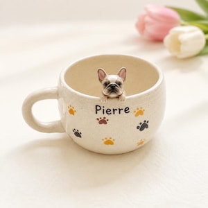 Könnte beinhalten: Ein cremefarbener Keramikbecher mit gesprenkelter Oberfläche und einer kleinen 3D-Figur einer französischen Bulldogge, die über den Rand schaut. Die Außenseite ist mit bunten Pfotenabdrücken und dem Namen Pierre in schwarzer Schrift verziert.