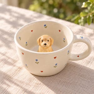 Può includere: Una tazza in ceramica color crema con una piccola statuetta tridimensionale di golden retriever sul fondo. L'esterno e l'interno sono decorati con un motivo di minuscoli fiori blu e piccoli cuori rossi, presente anche sul manico.