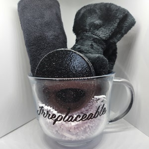 May include: A clear glass mug with a black glitter rim and a black fuzzy boot and a black fuzzy headband inside. The mug has the word "Irreplaceable" written on it in black.