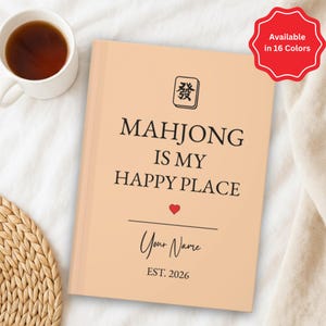 May include: A peach-colored notebook with the text "MAHJONG IS MY HAPPY PLACE" and a red heart. The notebook also includes the text "Your Name" and "EST. 2026". A red tag indicates the item is available in 16 colors. A cup of tea sits to the left.