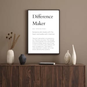 Puede incluir: Un marco negro con la inscripción "Difference Maker". La impresión incluye una definición y un texto inspirador. La impresión se exhibe en un mueble de madera con jarrones decorativos.