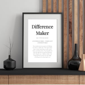 May include: Framed print with the words "Difference Maker" in a bold, modern font. The print includes a definition and inspirational text about making a difference. The black frame sits on a dark surface.