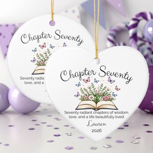 May include: Two white ceramic ornaments, one round and one heart-shaped, featuring the text Chapter Seventy, an open book with flowers, and butterflies. The heart ornament includes the name Lauren and the year 2026 with gold hanging strings.