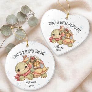 May include: Two white ceramic ornaments, one heart-shaped and one circular, each featuring a cartoon turtle with a house on its shell, a red scarf, and a wreath. The text reads Home Is Wherever You Are, with the names Jason or Rebecca and 2026.