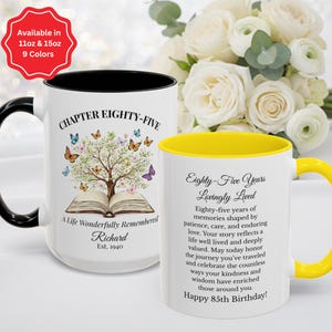 May include: Two ceramic mugs with white exteriors. The left mug has a black handle and rim, featuring a tree growing from an open book with butterflies and the text 'CHAPTER EIGHTY-FIVE A Life Wonderfully Remembered Richard Est. 1940'. The right mug has a yellow handle and rim, featuring the text 'Eighty-Five Years Lovingly Lived' followed by a paragraph about memories and 'Happy 85th Birthday!'.