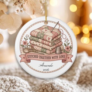 May include: A round white ceramic ornament featuring a stack of patterned quilts, a needle with thread, scissors, and a thimble. A pink banner displays the text STITCHED TOGETHER WITH LOVE, with the name Amanda and the year 2026 printed below.