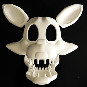 May include: A white, stylized mask of a fox-like character with large ears, wide eye openings, and a detailed jawline with sharp teeth. The mask is made of a solid, opaque material and is set against a black background.