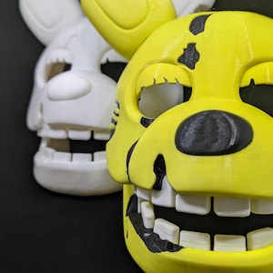 May include: Two animatronic masks, one white and one yellow with black details. The yellow mask has a wide smile with white teeth and black eyes.
