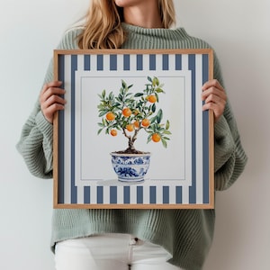 Peut inclure: Une impression encadrée d'aquarelle d'un oranger dans un pot décoratif bleu et blanc. L'œuvre présente une bordure à rayures bleues et blanches et un cadre en bois naturel. L'arbre est chargé d'oranges mûres.