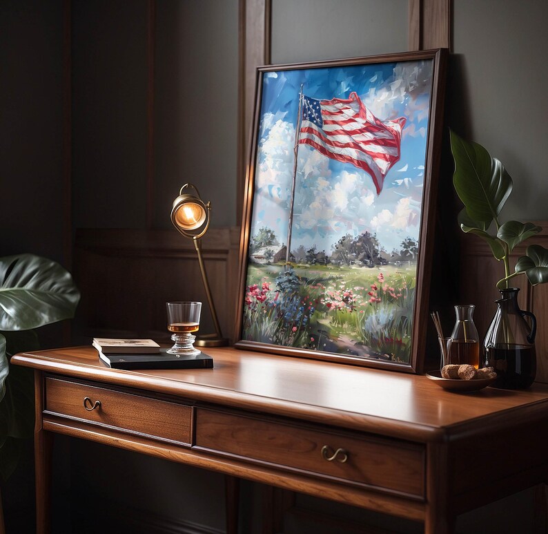 May include: A framed painting of the American flag waving in the wind, set against a blue sky with fluffy clouds. The artwork is displayed on a wooden desk with a lamp, a glass of amber liquid, and a small plant.