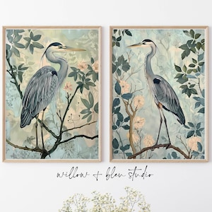 Op de afbeelding: Twee ingelijste prints van een blauwe reiger die op een tak staat in een blauwe en groene bloemenomgeving. De prints zijn in een licht houten lijst.