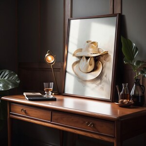 May include: A framed painting of two beige cowboy hats stacked on top of each other, with a light brown background. The painting is hanging on a wall above a wooden desk with two drawers.