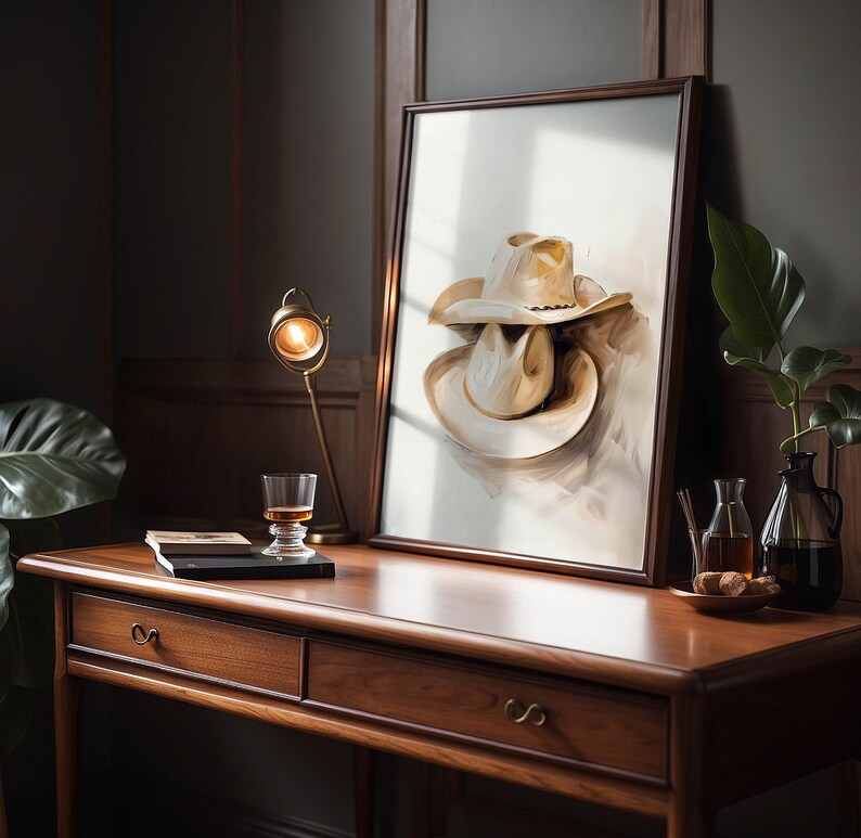 May include: A framed painting of two beige cowboy hats stacked on top of each other, with a light brown background. The painting is hanging on a wall above a wooden desk with two drawers.