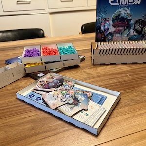 May include: Wooden board game organizer with various components. Includes card holders, trays with colored game pieces, and a box with game cards. The game box is visible in the background. The components are arranged on a wooden table.