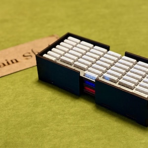 Può includere: Una scatola di legno marrone scuro con più scomparti pieni di piccoli rettangoli bianchi. La scatola ha un'etichetta con la scritta "Train St". Lo sfondo è verde.