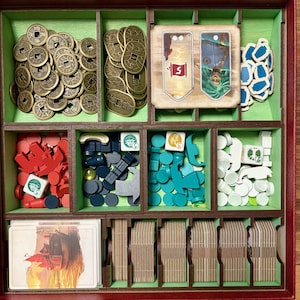 May include: A wooden game organiser with various compartments containing game pieces. These include gold coins, red and blue tokens, dice, and cards. The organiser is brown with green and red accents.