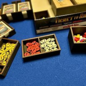 May include: A set of wooden game organizers for the board game Ticket to Ride. The organizers are divided into compartments to hold the game's various components, including train cars, destination tickets, and colored train markers.