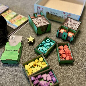 May include: A green wooden box with the text "Scoring Cards" on the front. The box is filled with small cards. There are also several other wooden boxes filled with colorful game pieces. The boxes are on a gray surface.