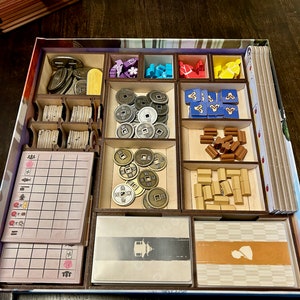 Könnte beinhalten: Ein hölzerner Organizer-Einsatz für ein Brettspiel mit Fächern für verschiedene Spielsteine, darunter Münzen, Kacheln und Holzsteine. Der Einsatz ist aus hellem braunem Holz gefertigt und hat ein blaues und weißes Design.