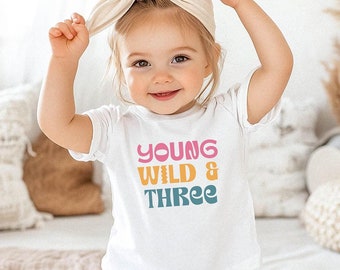 以下が含まれることがあります： カラフルなレトロなフォントで「YOUNG WILD & THREE」と書かれた白いTシャツ。薄い茶色のヘッドバンドをした小さな子供が着用しています。