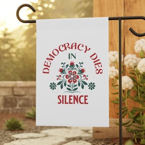 Könnte beinhalten: Weiße Gartenflagge mit einem roten und grünen Blumendesign und dem Text "Democracy Dies in Silence".
