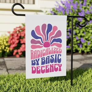 Op de afbeelding: Een witte tuinvaandel met een retro bloemenontwerp in roze, paarse en blauwe tinten. De vlag toont de tekst "RADICALIZED BY BASIC DECENCY" in een speels lettertype. De vlag staat op een zwarte metalen standaard in een tuin.