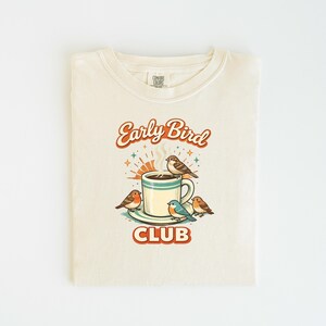 Könnte beinhalten: Gefaltetes cremefarbenes T-Shirt mit Retro-Grafik. Das Design zeigt eine Kaffeetasse mit Dampf, umgeben von Vögeln, und die Worte "Early Bird Club" in Orange und Braun. Das T-Shirt ist sauber auf einer weißen Oberfläche gefaltet.