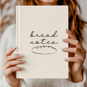 Könnte beinhalten: Ein cremefarbenes Notizbuch mit den Worten "bread notes" in handschriftlicher Schrift und einer Brotlaib-Illustration. Das Notizbuch wird vor einer Person hochgehalten.