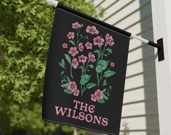 Może przedstawiać: Czarna flaga z różowymi kwiatami i zielonymi liśćmi. Tekst "THE WILSONS" jest nadrukowany na fladze kolorem różowym.