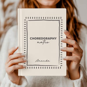 May include: A cream-colored hardcover notebook featuring a dark gray hand-drawn rectangular border with dashed lines. The text CHOREOGRAPHY notes is printed in the center, with the name Amanda written in a cursive script font below it.