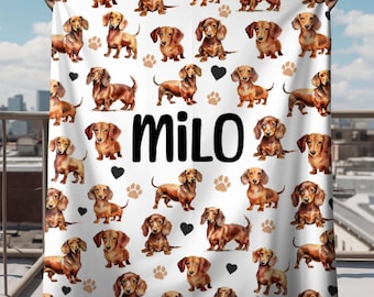 Puede incluir: Manta blanca con un patrón repetido de perros salchicha marrones, huellas de patas y corazones. La manta tiene el nombre "MILO" impreso en letras negras.