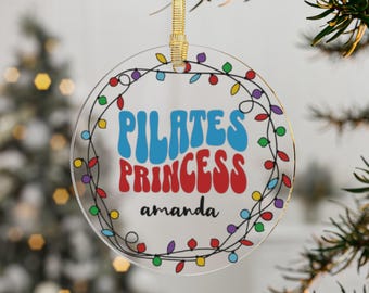 Könnte beinhalten: Ein runder, transparenter Glas-Anhänger mit einer bunten Lichterkette am Rand. Der Anhänger hat den Text "Pilates Princess" in einer Retro-Schriftart mit dem Namen "amanda" darunter.