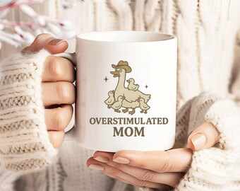 Peut inclure: Une tasse à café en céramique blanche avec une illustration de dessin animé d'une oie portant un chapeau de cowboy et entourée de petits oisons. Le texte "Overstimulated Mom" est imprimé sur la tasse.
