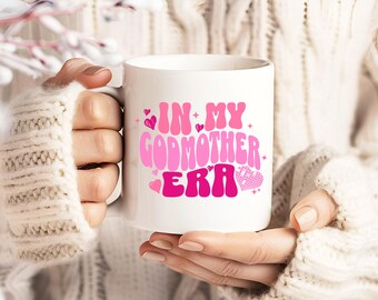May include: White ceramic mug with a pink and white design that says "In My Godmother Era" with pink hearts and stars.
