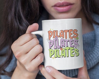 May include: White ceramic mug with a colorful design that says "PILATES" three times. The mug is decorated with flowers, stars, and smiley faces.